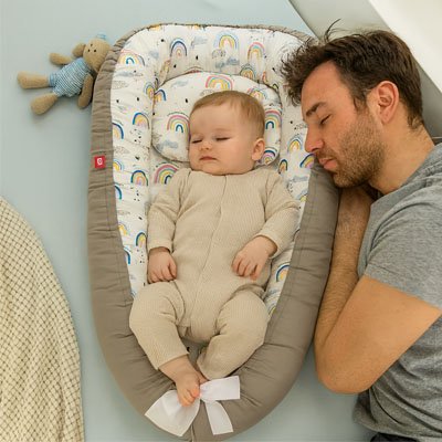 Père endormi et détendu à côté de son bébé qui dort en toute sécurité dans le [Cocon'Dodo™ / Réducteur de Lit Cocon], illustrant les nuits paisibles obtenues grâce au cododo sécurisé.