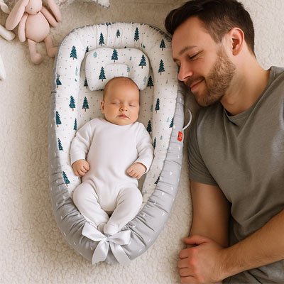 Père souriant et détendu à côté de son bébé joyeux installé confortablement dans le [Cocon'Dodo™ / Réducteur de Lit Bébé], soulignant la sérénité parentale et le bien-être de l'enfant.