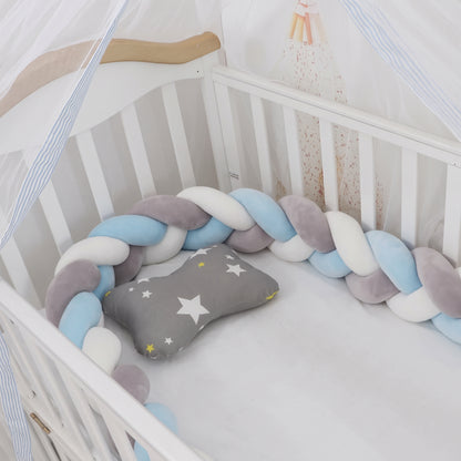 Tour de lit bébé tressé 3 brins aux couleurs bleu ciel, gris clair et blanc, installé dans un berceau en bois blanc avec un coussin en forme d'os à motif étoiles.